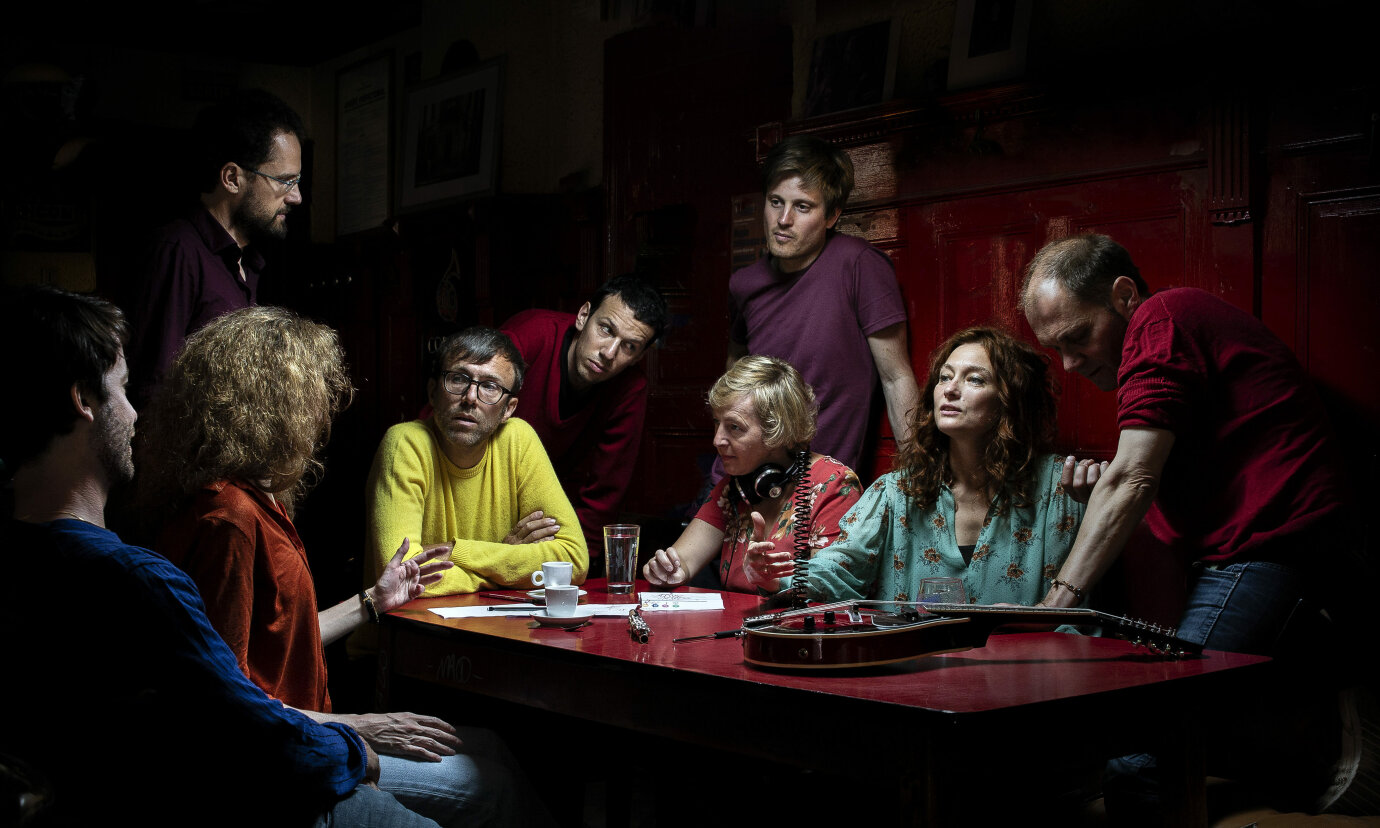 Bild des Ensembles in Residence Ictus: Die 9 Musiker*innen sitzen und stehen zusammen um einen Tisch und unterhalten sich angeregt.