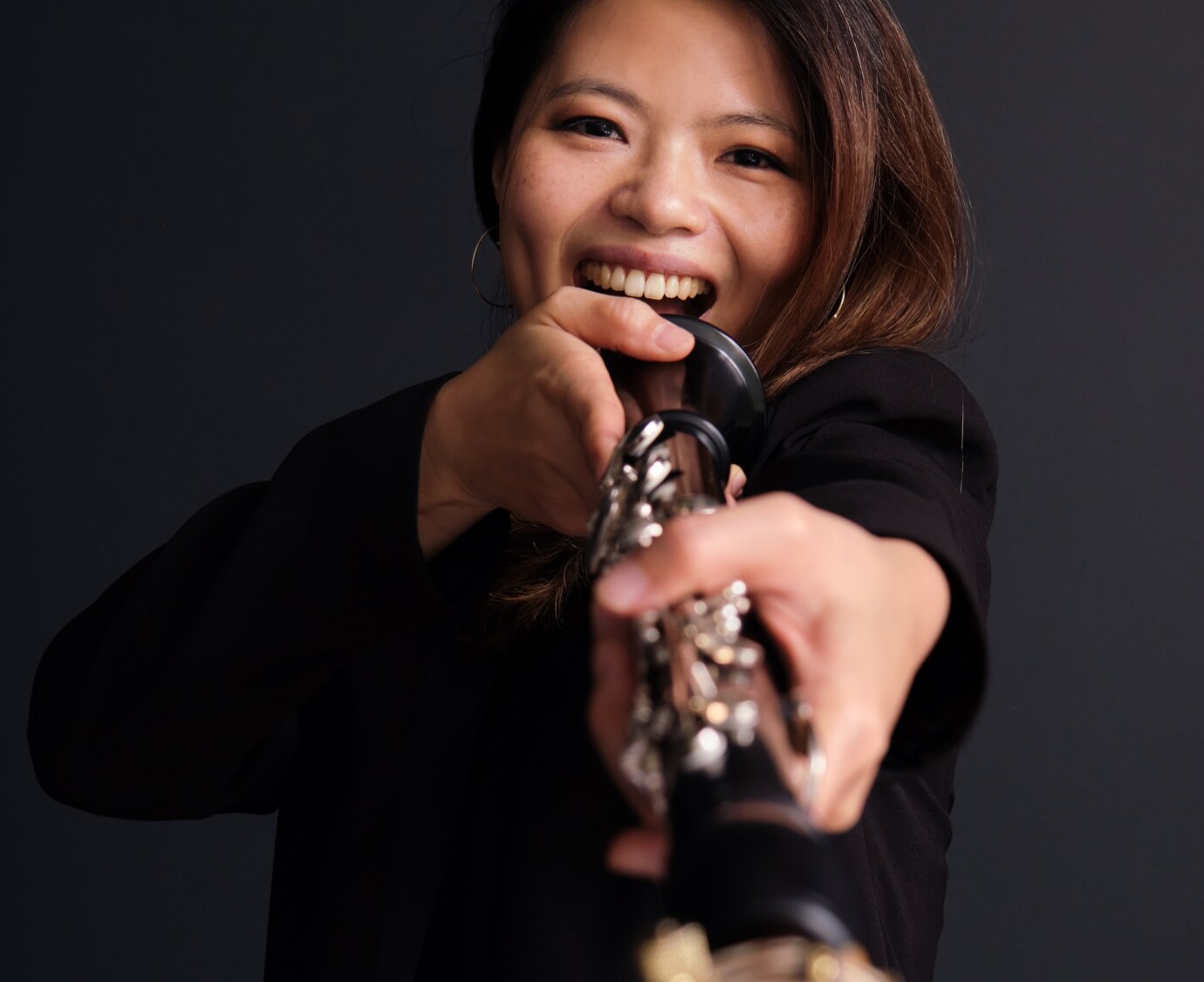 Die Klarinettistin Irene Chen hält ihr Instrument mit dem Mundstück nach vorne Richtung Kamera. Das Instrument ist vorne unscharf; das Porträt der lachenden Musikerin ist klar erkennbar. 