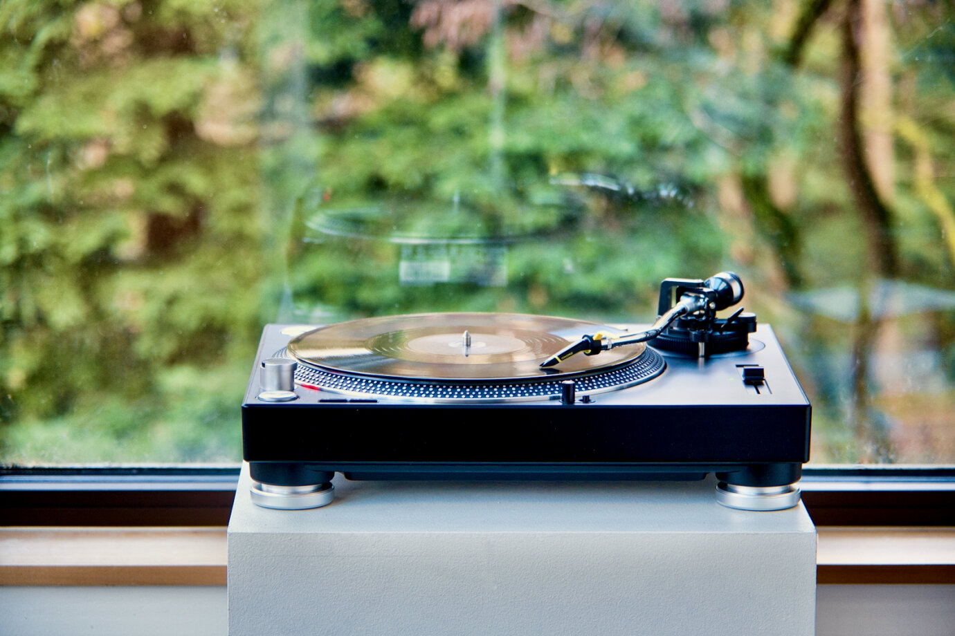 Ein Plattenspieler mit einer Vinylplatte steht auf einem weissen Podest. Im Hintergrund ist ein grosses Fenster. Der Plattenspieler spiegelt sich leicht darin. Das Fenster blickt in die Natur. Die Sicht nach aussen ist verschwommen.

